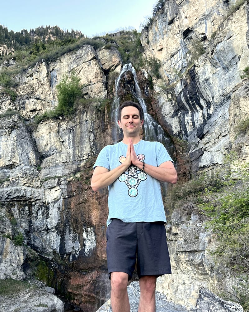 Jess standing at the foot of a waterfall, palms together at his chest, eyes closed.