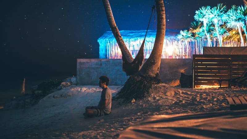 Jess sitting cross-legged on the sand under a palm at night, stars over a lit beach hut.
